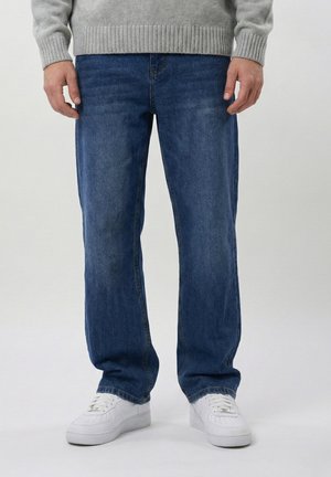 Mann iført rettbeinte blå jeans, grå genser og hvite joggesko, stående på en enkel lys bakgrunn.