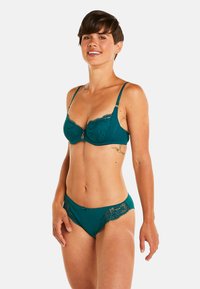 Ensemble de lingerie en dentelle teal comprenant un soutien-gorge et un slip assorti, mettant en avant des finitions en dentelle délicate, des bretelles ajustables et un petit nœud accentué sur le slip. Texture lisse.
