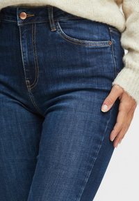 Tummansininen farkkushortsit, joissa on korkea vyötärö. Ne ovat varustettu kuparinvärisellä napilla, oranssilla ompelella ja sileällä pinnalla, kuvattuna käsi reisillä.