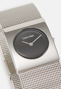 Montre carrée en argent avec un cadran noir, aiguilles des heures et des minutes argentées, et bracelet en maille métallique. Nom de la marque "Calvin Klein" affiché sur le cadran.