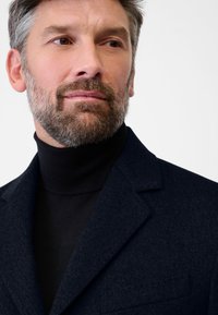 Dunkelblauer Wollmischblazer mit Reverskragen, strukturiertem Stoff und einer Brusttasche, getragen über einem schwarzen Rollkragenpullover. Gut definierter Kragen.