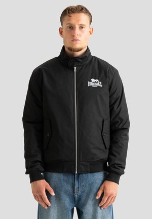 Homme portant une veste noire zippée Lonsdale avec un col haut et un jean bleu, debout devant un fond clair uni.
