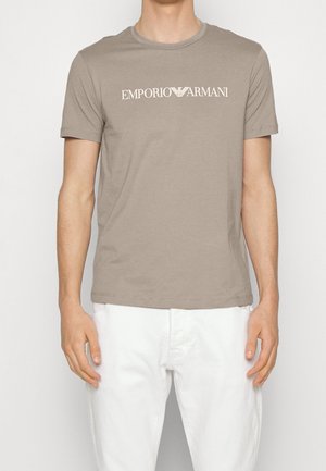 Personne portant une chemise à manches courtes taupe Emporio Armani avec un logo blanc, associée à un pantalon blanc, debout devant un fond uni.