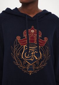Sweat à capuche en tricot bleu marine avec cordon de serrage, présentant un emblème brodé complexe en or et bordeaux ainsi qu'un motif de couronne sur la poitrine.