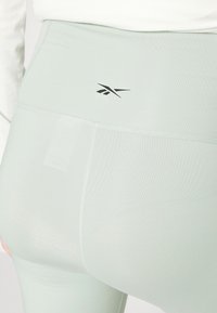 Leggings verde claro com um cós alto, tecido elástico e um pequeno logo preto na parte de trás. Textura suave com um brilho sutil.