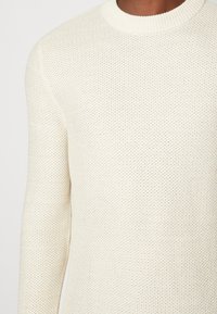 Krémově zbarvený pletený svetr s texturovaným vzorem, kulatým výstřihem a žebrovanými manžetami, který představuje bezšvový design a středně těžkou látku.