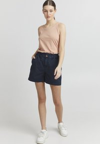 Top de tirantes sin mangas de color rosa claro combinado con pantalones cortos azul marino que tienen un botón frontal y una cinturilla elástica. Modelo lleva zapatillas blancas.