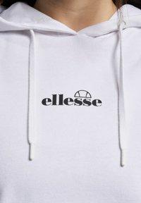 Nærbilde av hvit hettegenser med svart "ellesse" logo sentrert under halslinjen, med snorer på begge sider av hetten.