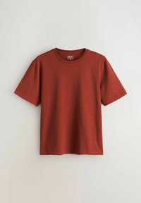 T-shirt en coton couleur rouille, manches courtes, col rond, taille moyenne, design uni, suspendu contre un mur clair.