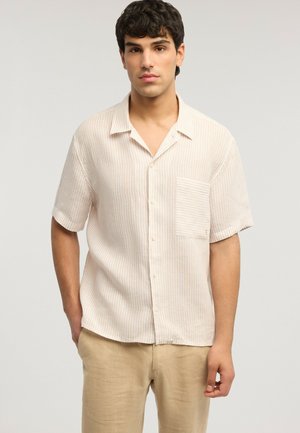 Ung mand iført en kortærmet, lysebeige, stribet skjorte med knapper og en brystlomme samt beige bukser, stående med hænderne i lommerne.