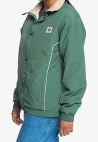 Zielona nylonowa kurtka z beżowym kołnierzem, zapinana na guziki z przodu, z białym przeszyciem. Na piersi logo oraz elastyczne mankiety.