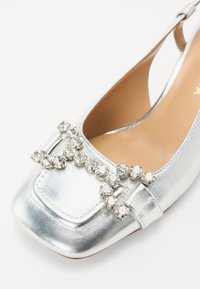 Silver leather slingback shoe with a square toe, adorned with a row of clear decorative crystals across the front. Smooth texture.