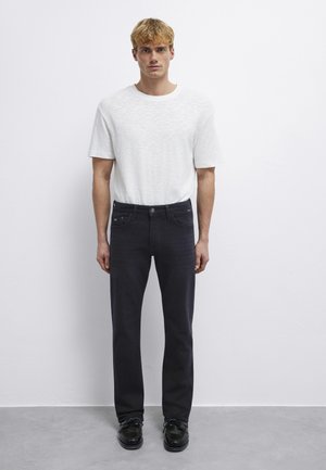 Junger Mann mit hellem Haar, der ein einfaches weißes Kurzarmhemd, schwarze Jeans und schwarze Schuhe trägt, steht vor einer weißen Wand.