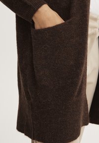 Cárdigan de lana marrón con un corte holgado, superficie texturada y un gran bolsillo en el lado izquierdo. Dobladillo ligeramente curvado.