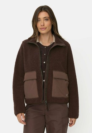 Kvinde iført en brun fleecejakke med store forlommer, lag-på-lag over en knappet sweater og en beige skjorte, stående foran en ensfarvet baggrund.