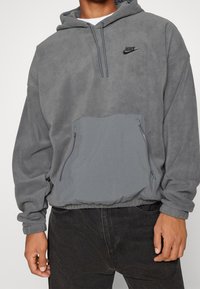 Šedá fleecová mikina s přední kapsou na zip. Má kapuci se stahovací šňůrkou a malé černé logo na hrudi. Elastické manžety a spodní lem.