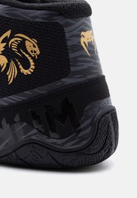 Πτέρνα μαύρου αθλητικού παπουτσιού με χρυσά λογότυπα φιδιού και αράχνης και υφασμένη λεπτομέρεια στη σόλα από καουτσούκ.