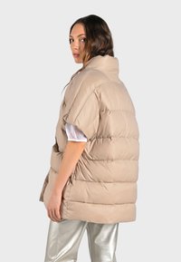 Beige pufferväst med hög krage, korta ärmar och horisontell quiltning. Bärs över en vit blus, som är kombinerad med silverfärgade metalliska byxor.