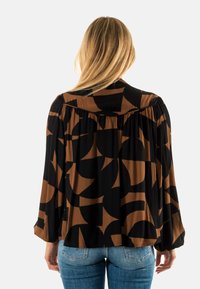 Blusa negra con patrón abstracto marrón, mangas largas, detalle fruncido en la parte trasera y ajuste relajado. Combinada con jeans de mezclilla azules.