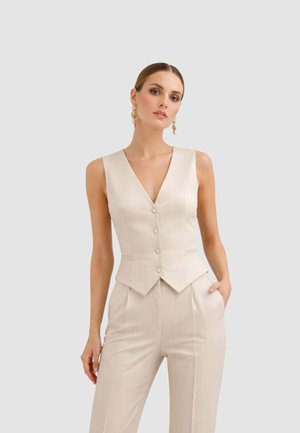 Donna che indossa un gilet senza maniche color crema con bottoni e pantaloni a vita alta abbinati, in piedi con una mano in tasca davanti a uno sfondo semplice.