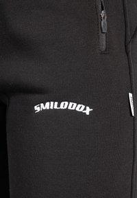 Svarta joggingsbyxor med en dragkedjeficka, slät textur och en vit logotyp som säger "SMILODOX" placerad nära midjan.