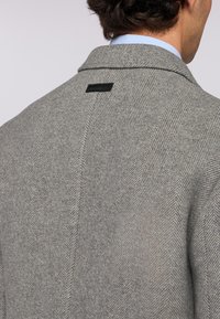 Šedý vlněný kabát s vzorem herringbone. Má klopový límec, jemnou texturu a malou černou značku na zadní straně.