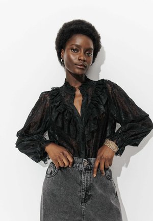 Blouse en dentelle noire avec des accents froncés, manches longues et col montant, assortie à un jean en denim gris taille haute.