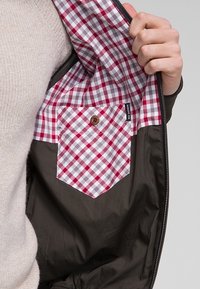 Ruka držící otevřenou tmavou bundu, která ukazuje červenou a bílou kostkovanou vnitřní kapsu s knoflíkovým uzávěrem a štítkem "Ben Sherman".