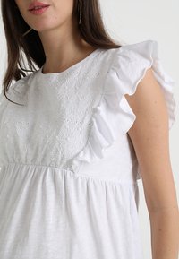 Top branco com um corpete texturizado, detalhes bordados, mangas com franzido e um corte relaxado. O tecido tem uma aparência suave e leve.