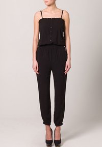 Combinaison noire à fines bretelles, avec un bouton à l'avant, une taille élastique et des jambes fuselées. Tissu lisse avec une coupe décontractée.