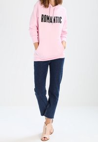 Sudadera con capucha de color rosa claro con "ROMÁNTICO" impreso en letras negras en negrita, combinada con jeans de color azul oscuro y botas de tobillo abiertas de color beige.