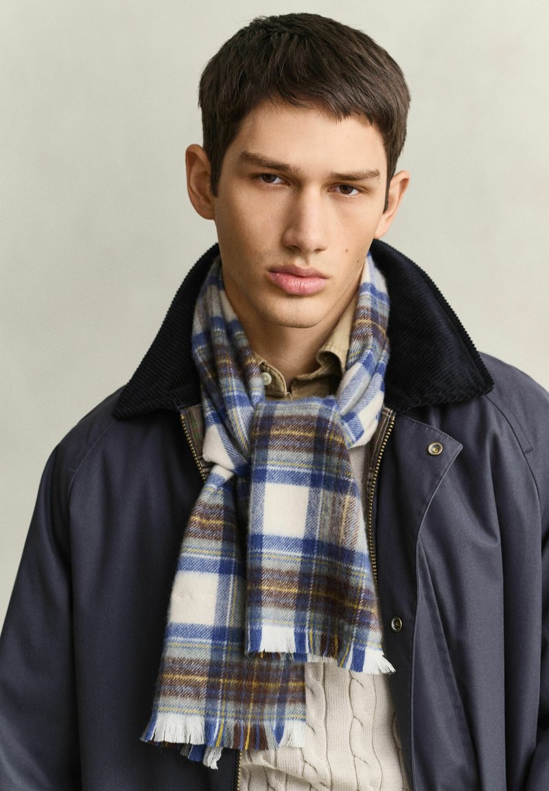 Jeune homme aux cheveux courts et foncés portant une veste bleue, un pull beige et une écharpe à carreaux bleue, blanche et marron sur un fond clair.