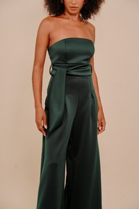 Sieviete, kas valkā stila bezplecu tumši zaļu satīna kombinezonu ar platām kājām un sānu drapējumu uz bēša fona.