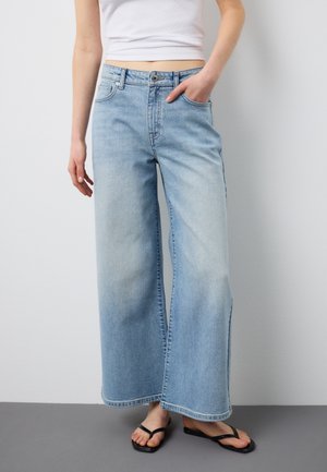 Persona che indossa jeans azzurri a gamba larga con vita alta, top bianco corto e infradito nere, in piedi contro un muro bianco.