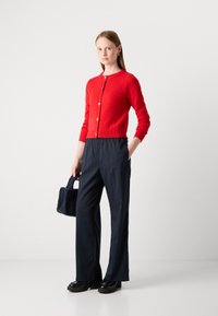 Červený pletený cardigan s knoflíkovým zapínáním, doplněný námořnickými pruhovanými kalhotami. Modelka drží tmavě modrou chlupatou kabelku. Silné černé boty.