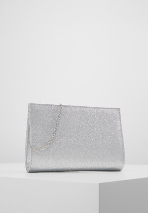 Pochette pailletée argentée avec une fine chaîne, posée sur une surface blanche angulaire devant un fond uni.