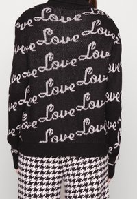 Černý pletený svetr s opakujícím se vzorem "Láska" v bílé barvě. Hladký límec a manžety, volnější střih. Nosený s kalhotami s kostkovaným vzorem.