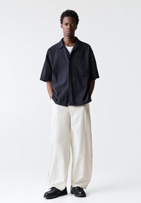 Short-sleeve black shirt with a chest pocket, paired with loose-fitting white trousers and black shoes. Smooth fabric and relaxed design.