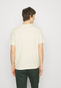 Kortærmet, lys beige t-shirt lavet af tekstureret stof; har en rund halsudskæring og et side-tag accent. Bæres med mørkegrønne bukser.