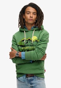 Groene hoodie met een grafisch bergontwerp, trekkoorden en geribde manchetten. Gedragen over een denimshirt, gecombineerd met een blauwe jeans.