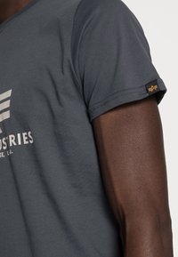 Dunkelgraues T-Shirt mit kurzen Ärmeln und Rundhalsausschnitt, ausgestattet mit einem weißen grafischen Druck und einem Markenetikett am Ärmel.