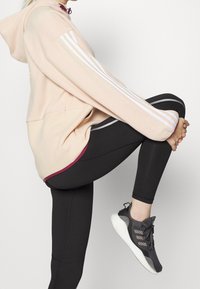 Světle růžová mikina s bílými pruhy na rukávech, černé legíny se šedými detaily a černé sportovní boty s texturovanými prvky.