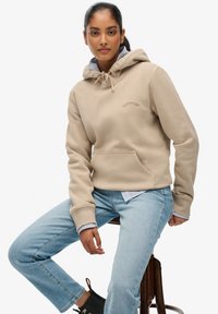 Beige hoodie met trekkoord, voorzak en geborduurd logo. Gecombineerd met lichtblauwe spijkerbroek en een gestreept overhemd eronder.