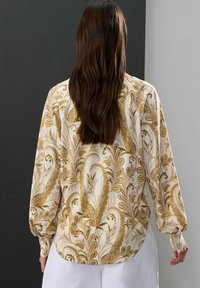 Blouse à manches longues en crème avec un motif paisley doré complexe, présentant une coupe décontractée et une texture douce et soyeuse.