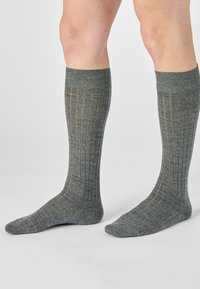Gambe che indossano calze a coste grigie fino al polpaccio, in piedi su una superficie chiara contro uno sfondo neutro.