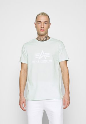 Lys mintgrøn bomulds t-shirt med korte ærmer, prydet med et hvidt logo og et enkelt design. Bæres med hvide bukser.