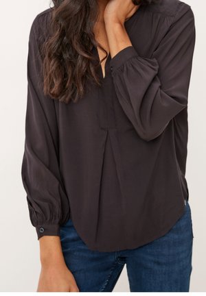 Femme portant un chemisier noir à manches longues avec des poignets froncés et un col en V, associé à un jean bleu, posant avec une main près du cou.