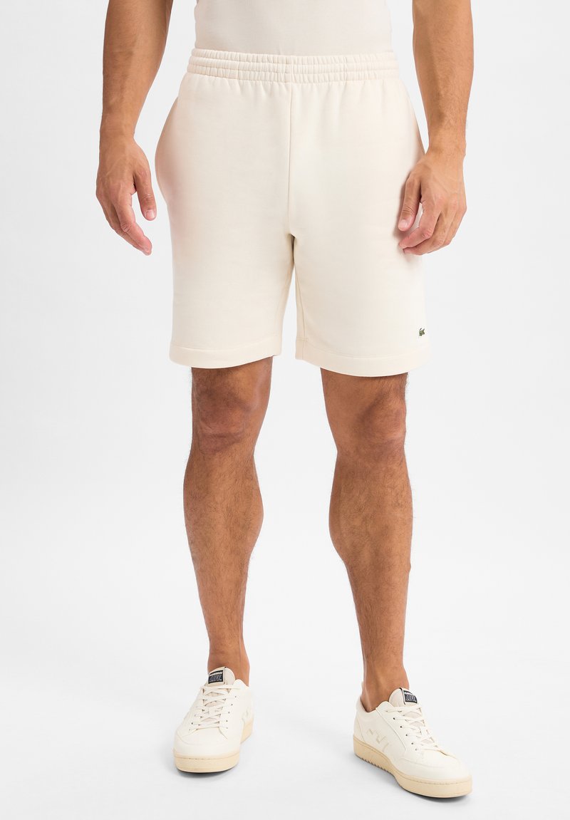 Roomkleurige shorts met een elastische tailleband, knielengte, zachte textuur en een klein groen logo linksonder.