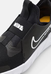Zapatilla deportiva negra con parte superior texturizada, correa elástica y logo de Nike blanco. Cuenta con un cuello acolchado y una suela blanca en contraste.