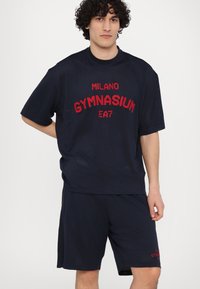 Navy tīkla sporta komplekts, kas sastāv no īsām piedurknēm krekla un šortiem. Krekls ir ar faktūru un uz tā ir sarkanski „MILANO GYMNASIUM EA7” teksts.